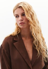 Hnědý, lehký blejzr s hlubokým V-výstřihem, klopovými límci a volným střihem. Tkanina má jemnou texturu a hladký povrch.