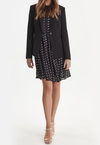 Czarna dopasowana marynarka na czarno-białej sukience w kropki, z falbaną u dołu i guzikami, w połączeniu ze złotymi sandałkami na paski.