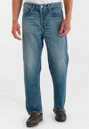 Man draagt vervaagde blauwe straight-leg jeans en donkerbruine leren veterschoenen, staat met ontspannen handen langs het lichaam.