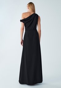 Czarna wieczorowa suknia na jedno ramię z drapowanym górnym fragmentem i dopasowaną talią, przechodząca w długą spódnicę z gładkiego materiału.