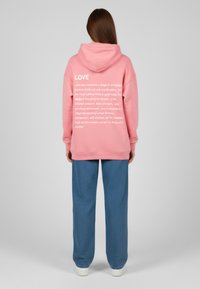 Felpa con cappuccio rosa con testo bianco sul retro, abbinata a pantaloni blu larghi e sneakers bianche, su uno sfondo neutro.