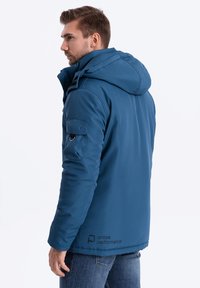 Modrá izolovaná bunda s kapucí, která má zipovou kapsu na rukávu, hladký povrch a vyšitý logo na dolní části.