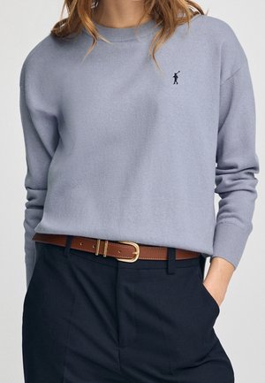 Kobieta ubrana w jasno niebieski sweter z okrągłym dekoltem z małym haftowanym logo gracza polo, brązowy pasek i czarne spodnie, z jedną ręką w kieszeni.