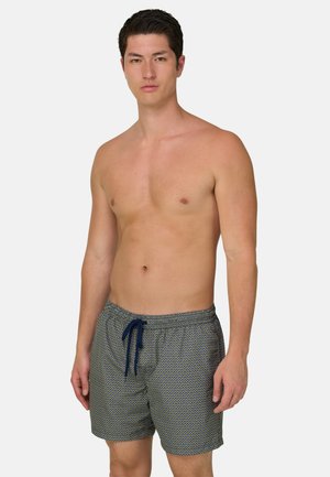 Joven con cabello corto y oscuro que lleva bañador con estampado y cordón azul marino, de pie frente a un fondo liso y claro.