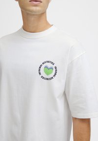 Hvid bomuld T-shirt med rund hals, der har et cirkulært grafisk design i grøn, blå og sort tekst på venstre brystområde.