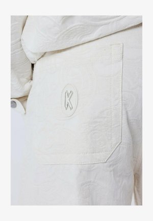 Tissu blanc avec un motif floral en relief et un patch rectangulaire comportant un logo "K" découpé sur la poche arrière. Texture lisse, forme ajustée.