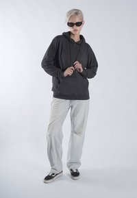 Sweat-shirt gris foncé avec capuche, coupe décontractée, poche avant, porté avec un jean clair déchiré et des baskets noires. Les accessoires comprennent des lunettes de soleil.