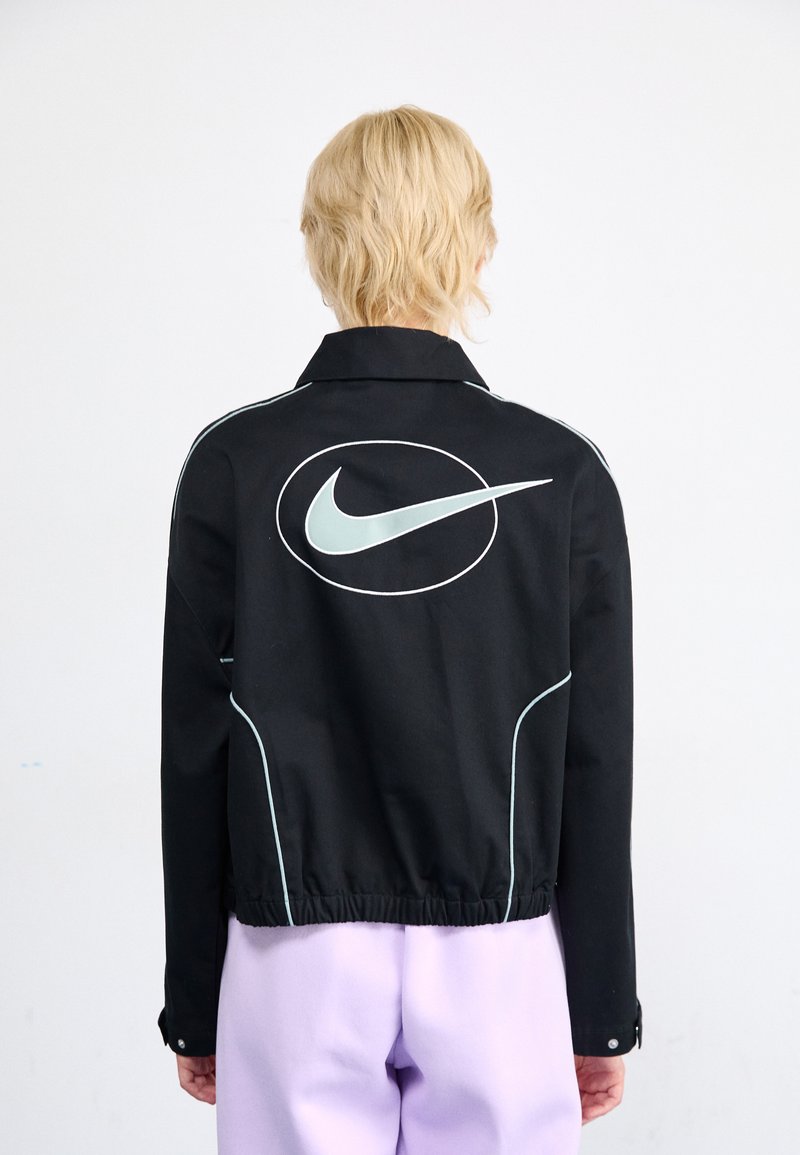 Chaqueta negra con un gran logo de Nike en blanco y azul claro en la parte posterior, con costuras en azul claro en contraste y un diseño con cuello.
