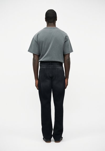 Άνδρας που στέκεται με την πλάτη, φοράει ένα ξεθωριασμένο γκρι μεγάλο T-shirt και φαρδιά μαύρα τζιν μπροστά σε λευκό απλό φόντο.