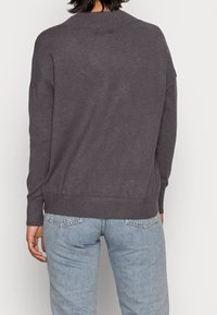 Dunkelgrauer Pullover mit geripptem Rundhalsausschnitt und überschnittenen Schultern. Weiche Textur, lockere Passform, kombiniert mit hellblauen Jeans.