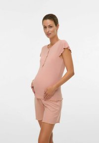 Femme enceinte portant un haut à manches courtes pêche et un short assorti, tenant doucement son ventre, debout devant un fond uni.