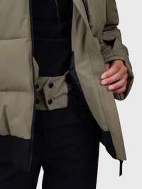 Pessoa a usar uma jaqueta verde com forro interior preto, calças pretas, e a segurar a jaqueta aberta para mostrar os botões de pressão no flap interior.
