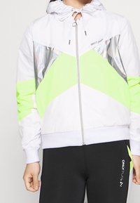Chaqueta blanca con cremallera y detalles en verde neón, además de paneles reflectantes en plata. Presenta una capucha y un ajuste estructurado. Textura suave.