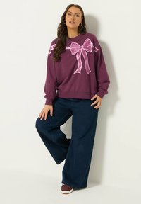 Kobieta stoi oparta o ścianę, ubrana w fioletowy sweter z dużym różowym nadrukiem w kształcie kokardy, szerokie granatowe dżinsy oraz fioletowe sneakersy.