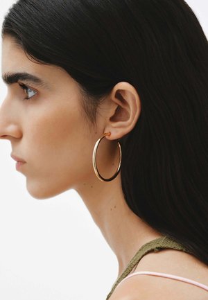 Pendiente de aro de metal dorado llevado en la oreja de un modelo, con una superficie lisa y brillante. El modelo tiene el cabello largo y oscuro y lleva una blusa verde.