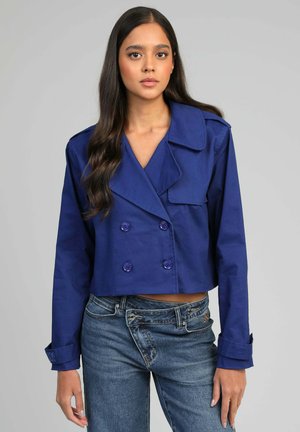 Jeune femme aux longs cheveux noirs portant une veste bleue croisée raccourcie et un jean bleu taille haute, debout devant un fond gris.