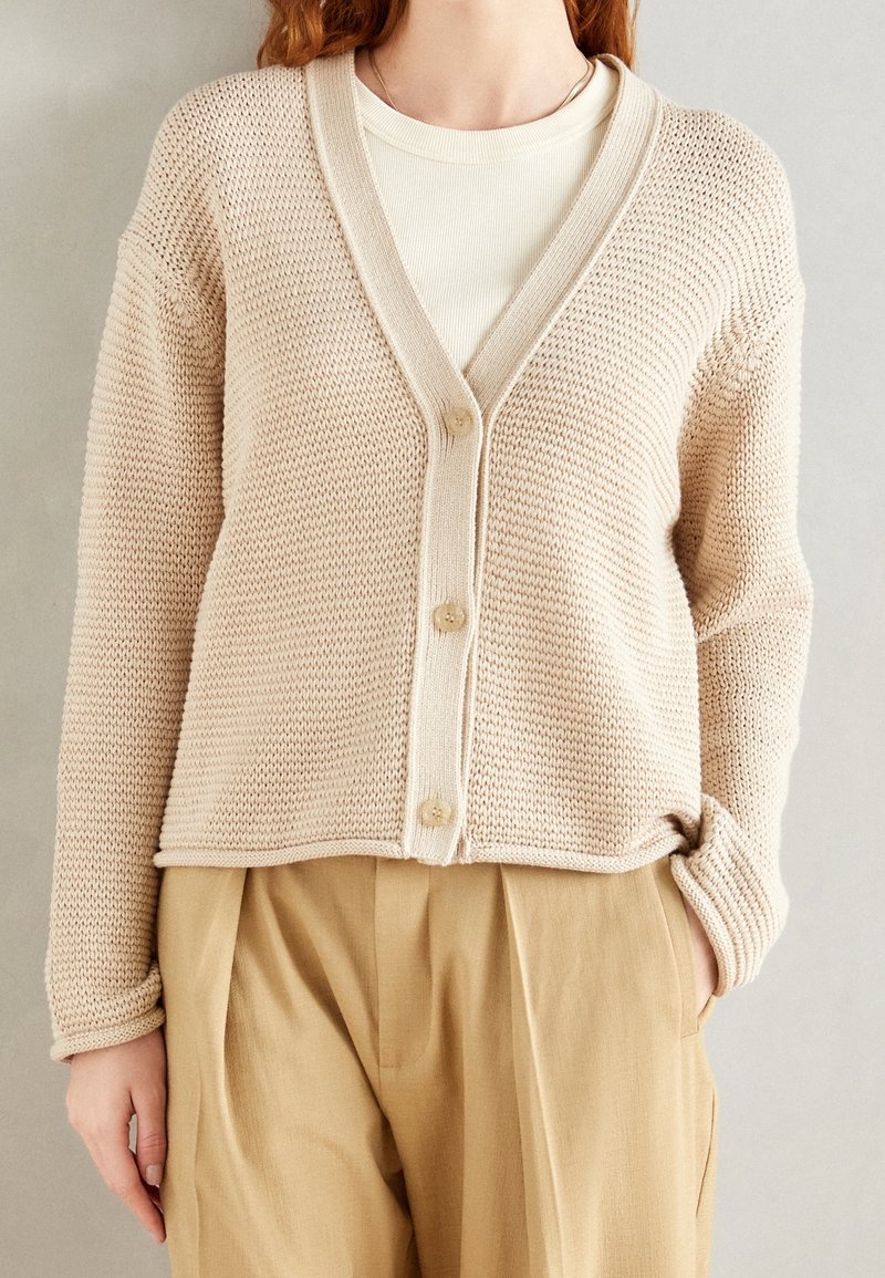 Cardigan beige lavorato a maglia con scollo a V, quattro bottoni e polsini a coste. Abbinato a pantaloni beige chiaro a vita alta, con una consistenza morbida.