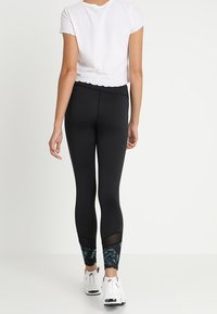 Černé legíny s síťovými panely a listovým vzorem na kotnících, zkombinované s bílým crop topem a bílými sportovními botami.