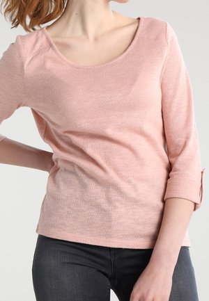 Femme portant un haut rose clair à manches longues avec des poignets retroussés et un pantalon gris foncé ajusté, sur un fond clair uni.