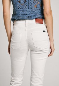 Hvite, tettsittende jeans med to baklommer, lærpatch på livet og glatt stofftekstur. En heklet blå topp er synlig.