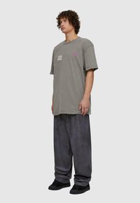 Pārlieku liels pelēks kokvilnas T-krekls ar drukātu tekstu, kombinēts ar brīvām melnām biksēm un melniem sneakers. Audums ir mīksts, nedaudz nolietots.