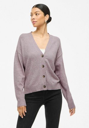 Frau trägt eine hellviolette, knöpfbare Strickjacke über einem weißen Oberteil mit schwarzen Hosen und schaut nach links vor einem schlichten Hintergrund.