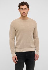 Beżowy sweter dziany z okrągłym dekoltem, długimi rękawami i ribbrowymi mankietami. Materiał ma teksturowany wzór. Noszony z czarnymi dżinsami.