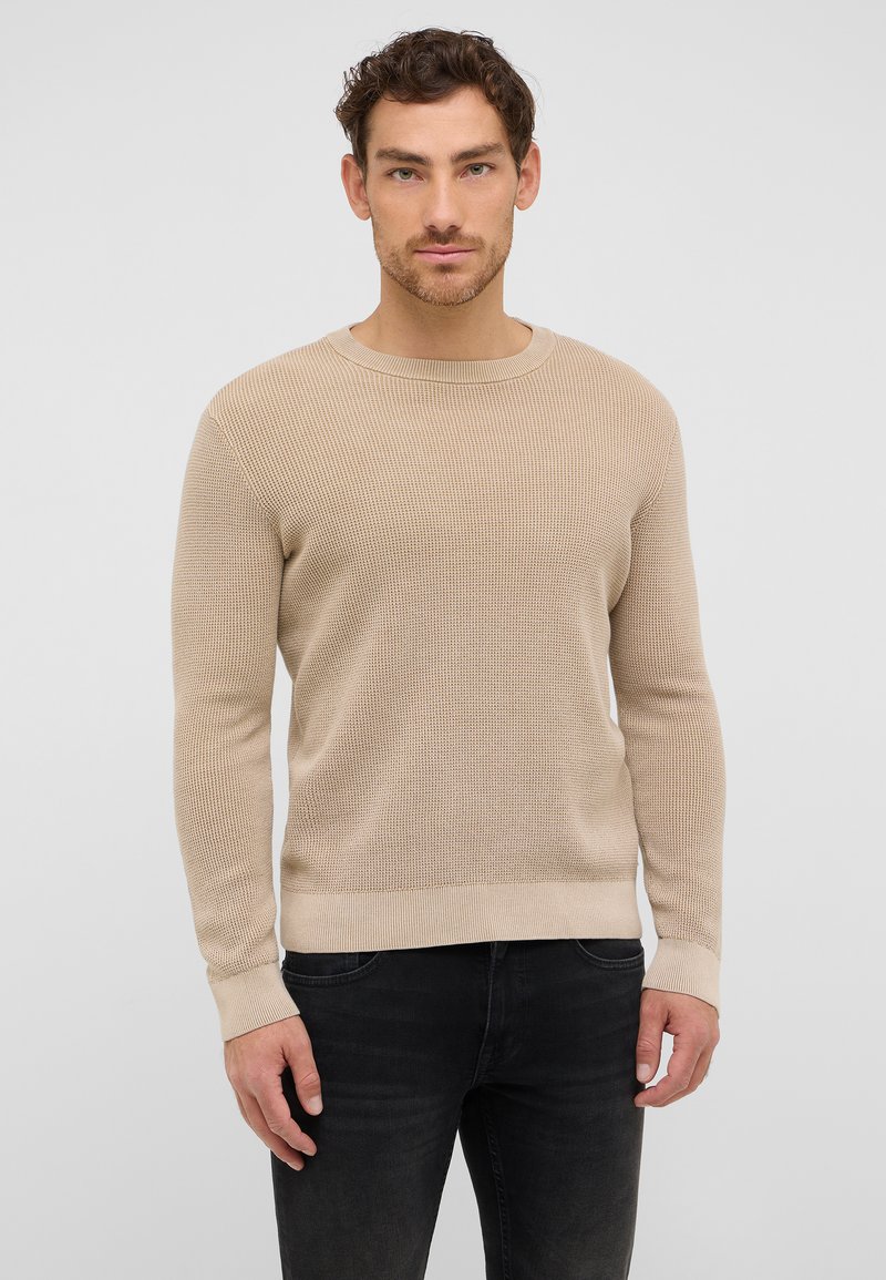 Beżowy sweter dziany z okrągłym dekoltem, długimi rękawami i ribbrowymi mankietami. Materiał ma teksturowany wzór. Noszony z czarnymi dżinsami.