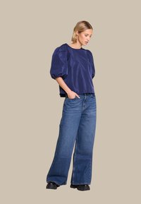 Marineblå, satinaktig topp med pufferm, kombinert med lyseblå denimjeans med vide ben og kraftige svarte støvler. Enkel, minimalistisk design.