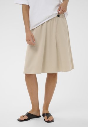 Osoba oblečená v béžovej sukni po kolená s elastickým pásom, bielom topu a čiernych otvorených sandáloch na plochej podrážke na jednofarebnom pozadí.