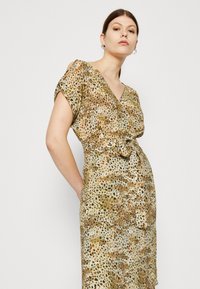 Leopardenmuster-Kleid mit kurzen Ärmeln, V-Ausschnitt und einem geknoteten Taillendetail. Der Stoff zeigt eine Mischung aus Brauntönen und Grüntönen in einem leichten, strukturierten Material.