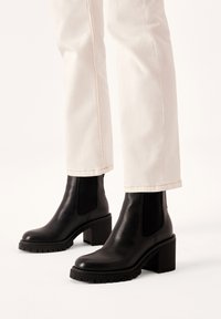 Czarne skórzane botki za kostkę z grubym obcasem i elastycznymi panelami bocznymi. Teksturowana gumowa podeszwa z bieżnikiem dla lepszej przyczepności. W zestawie z jasnymi spodniami.