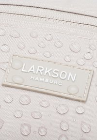 Svetlobéžová taška s textúrovanou tkaninou, s vodnými kvapkami a obdĺžnikovou etiketou s nápisom "LARKSON HAMBURG" bielym písmom.