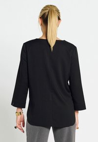 Maglietta nera a maniche lunghe con scollatura arrotondata, texture liscia e leggero orlo posteriore. Presenta un dettaglio di cucitura sul retro e aperture ai polsini discrete.