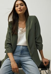 Olivov�ě zelený blazer s ohrnutými rukávy přes bílou kamizolu s krajkovým lemem, doplněný světle modrými džíny s vysokým pasem. Jednoduchý design, volný střih.
