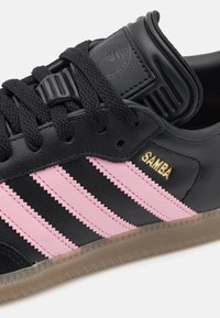 Svart lädersneaker med tre rosa ränder, texturerade accenter och en guld "SAMBA"-logga. Har svarta snörningar och en sula i gummigummi.