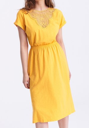 Femme portant une robe jaune longueur genoux avec manches retroussées et un détail en dentelle au niveau de l'encolure, debout sur un fond blanc.