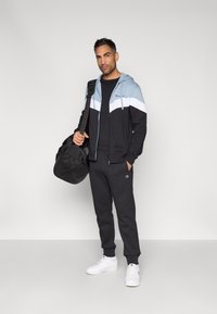 Homem vestindo um casaco com capuz preto e azul com fecho zip e calças de fato de treino pretas, segurando uma mala preta de ombro, em pé contra um fundo simples.