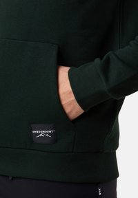 Felpa verde scuro con tasca a marsupio, polsini a coste e un'etichetta nera con la scritta "SWEDEMOUNT™" e grafiche di montagne in basso.