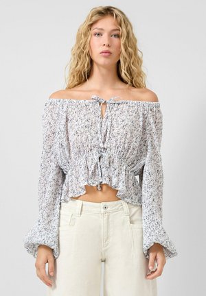 Blonde Frau trägt eine weiße, schulterfreie Bluse mit Blumenmuster, langen, aufgepufften Ärmeln und hellbeige, hoch taillierte Hose vor einfarbigem Hintergrund.