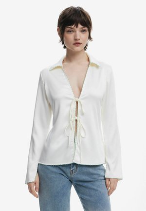 Blusa bianca a maniche lunghe con scollatura a V profonda, chiusura con fiocco davanti e bordi scallopati verde chiaro lungo il colletto e l'orlo.