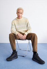 Pull en tricot jaune et gris avec une texture côtelée, associé à un pantalon en velours côtelé marron et des bottes de randonnée noires sur un sol bleu.