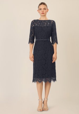Navyblaues Spitzenkleid mit Dreiviertelärmeln, welligem Saum und abnehmbarem Satinquas. Getragen mit silbernen Knöchelriemen-Absätzen.