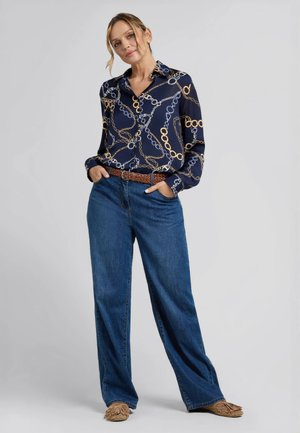 Donna in piedi con le mani nelle tasche, indossa una camicia blu navy con motivo a catena, jeans blu a gamba larga, cintura marrone intrecciata e mocassini color cuoio.
