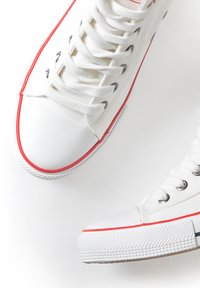 White canvas sneakers with red accents, flat laces, rubber toe cap, and textured outsole. Silver eyelets near laces are visible.