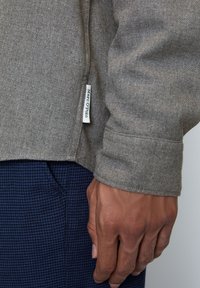 Gray textured fabric shirt with a visible cuff and brand tag; worn over dark patterned pants. Focus on shirt's material and design.