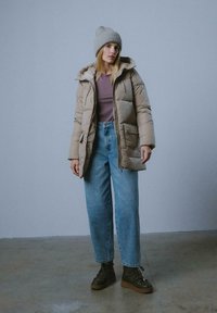 Beżowa kurtka puchowa z kapturem, z przodem na zamek i kieszeniami, noszona na fioletowej bluzce, w połączeniu z jasnoniebieskimi luźnymi dżinsami i zielonymi butami na sznurówki.