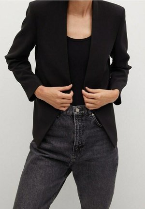 Person iført en sort blazer over en sort top og højtaljede mørkegrå jeans, holder blazerens revers med begge hænder.