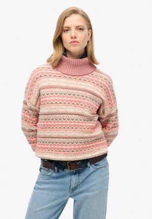 Rosa gemusterter Strickpullover mit hohem Rippkragen, der in einem geometrischen Design in Beige- und Grüntönen gestaltet ist, kombiniert mit hellblauen Jeans.