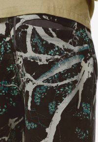Sort stof med et dristigt abstrakt mønster i teal og hvid, der viser blade og grene. Inkluderer en lille lomme detalje bagpå.